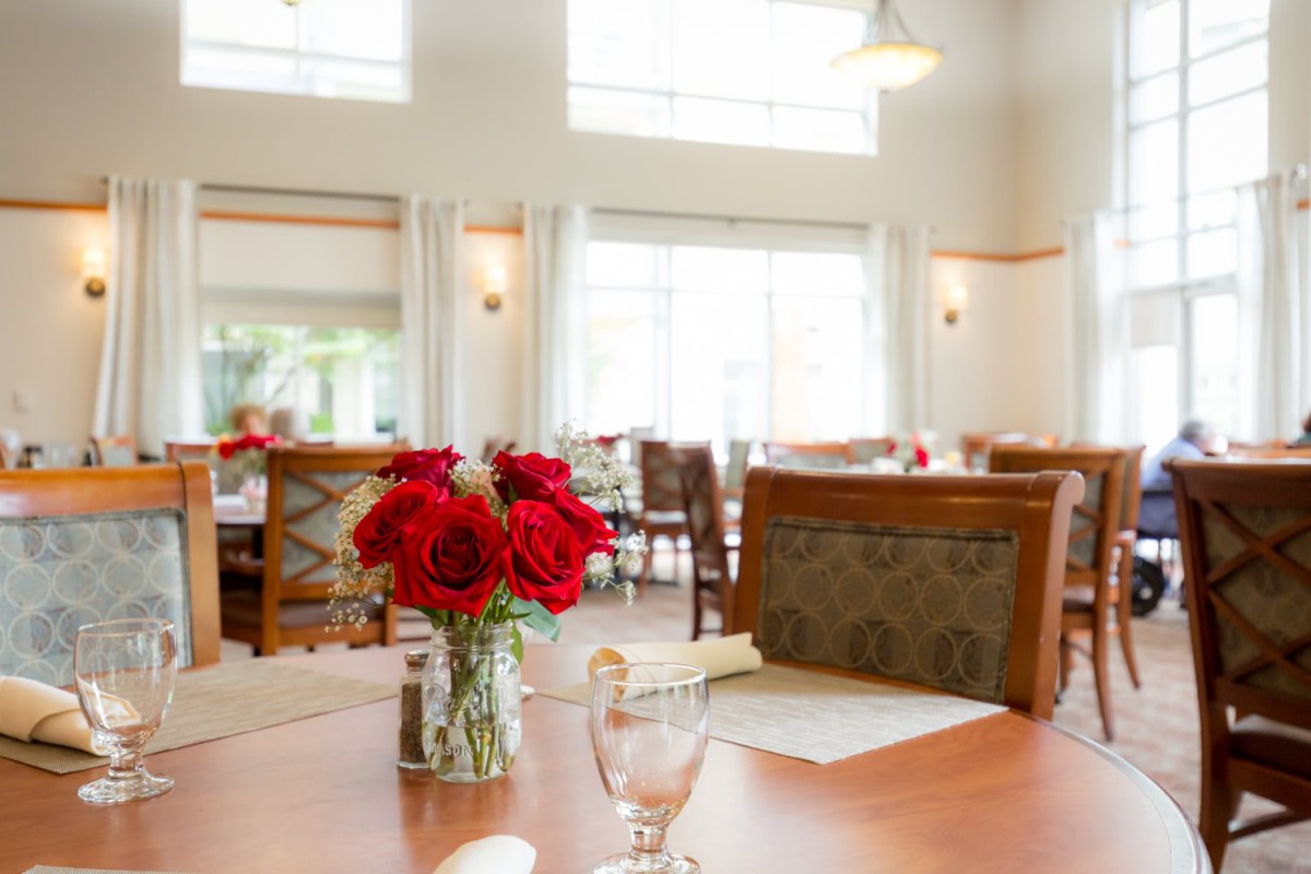 A well-lit dining area with tables set with red roses and chairs.