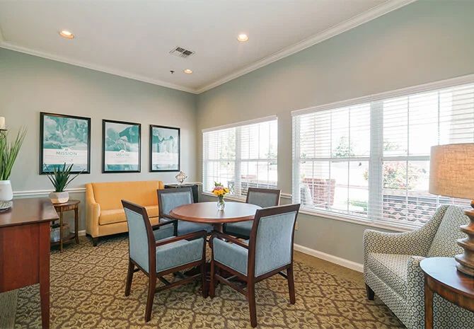 A cozy living room with chairs around a table, sofas, large windows, and decorative elements on the walls.