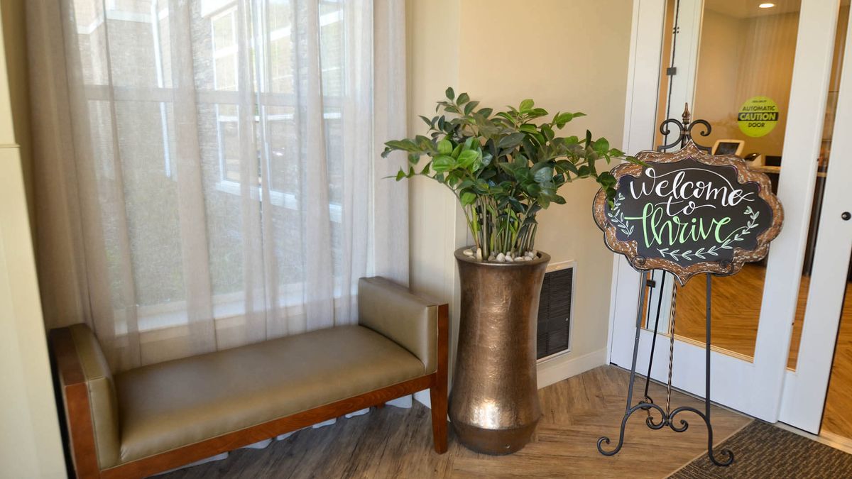 A cozy interior corner featuring a bench, large vase with greenery, and a welcome sign by the window.