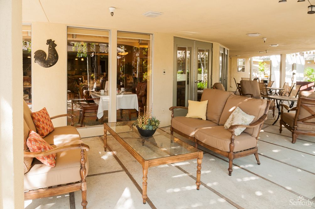 A cozy patio with comfortable outdoor furniture and a glass table, surrounded by lush plants.