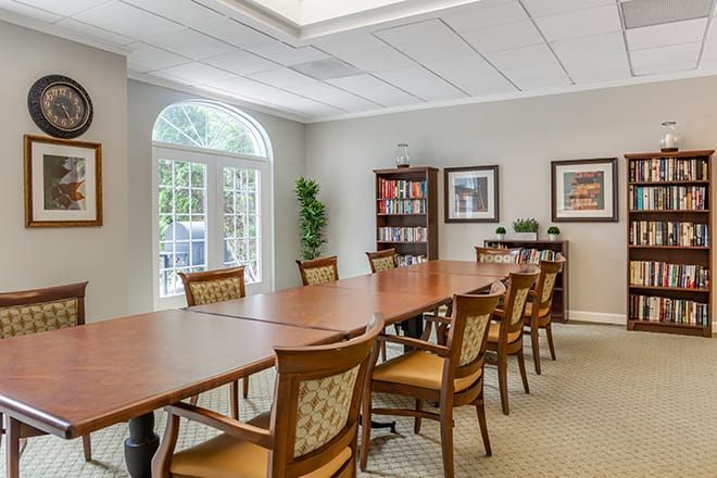 A well-lit conference room with a long wooden table and chairs, featuring a bookshelf and large window.