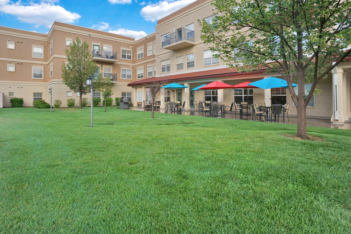 An outdoor view of a residential building with a spacious lawn and patio featuring colorful umbrellas and seating.