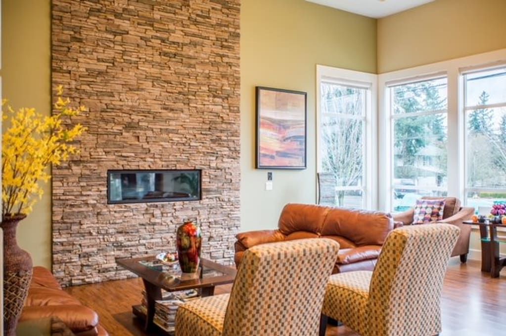 A cozy living room featuring a stone fireplace and modern furniture, illuminated by natural light from large windows.