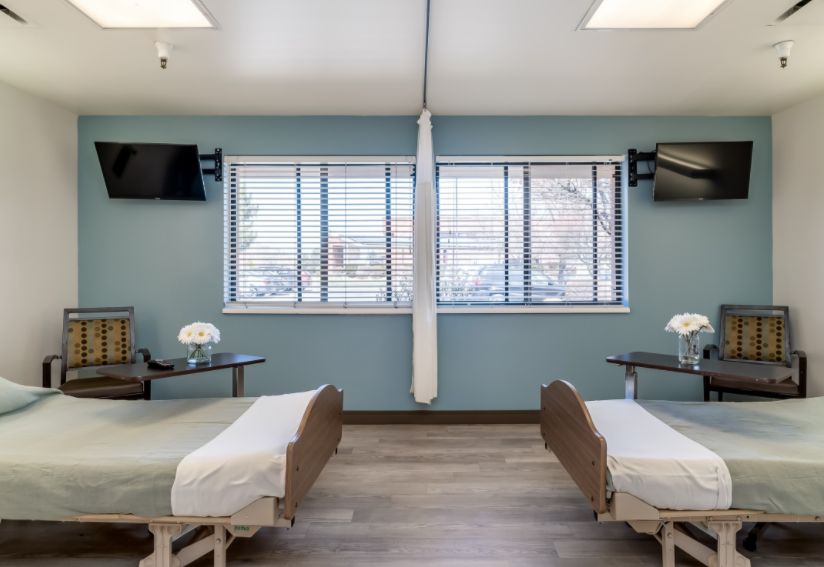 A modern hospital room with two medical beds separated by a curtain, featuring televisions and large windows.