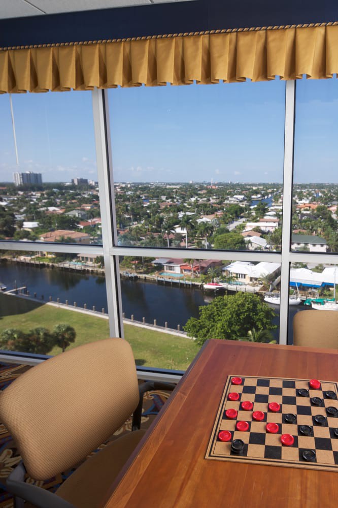 A room with a checkerboard game table and chairs overlooking a vibrant cityscape and waterfront.