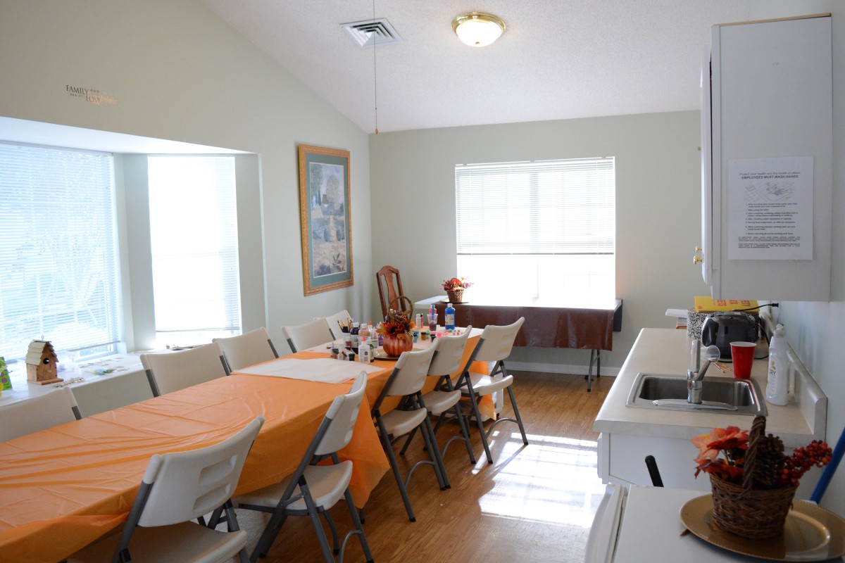 A dining room with a long table covered in an orange tablecloth, surrounded by folding chairs and lit by natural light from the windows.