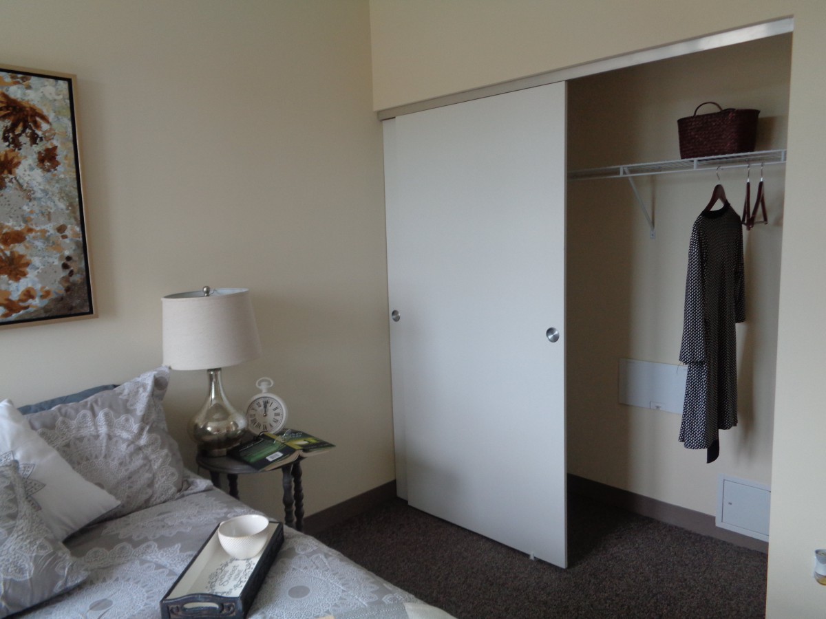 A bedroom with an open closet showing hanging clothes and a bedside table with a lamp.