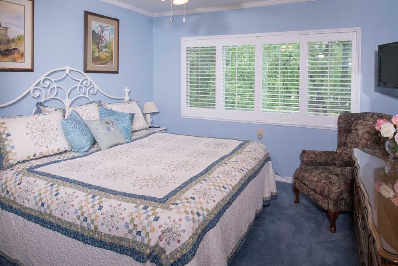 A cozy bedroom with soft blue walls, featuring a neatly made bed with a quilt and pillows. The room has a window with white blinds and a patterned chair in the corner.