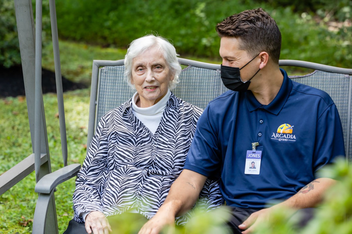 An elderly woman and a caregiver sit together outdoors, engaged in conversation.