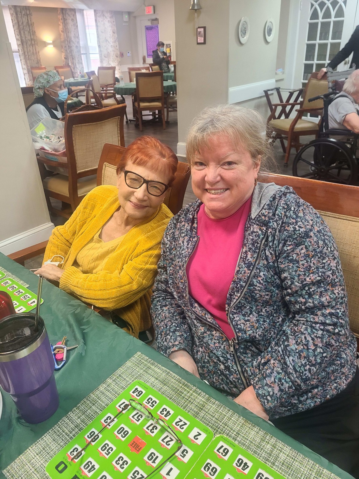 Two women are seated at a table, smiling in a social setting.