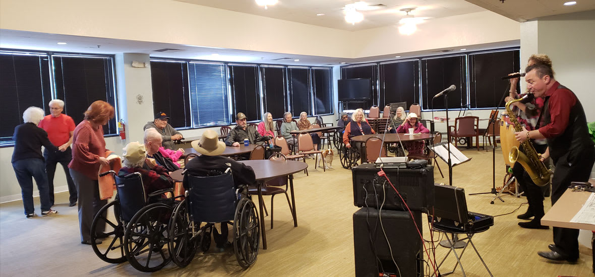 A community gathering of seniors in a meeting room, with several participants in wheelchairs.