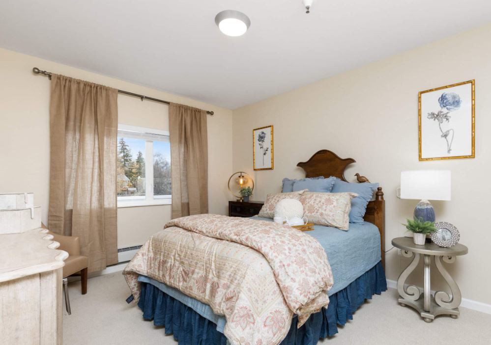 A cozy bedroom with a neatly made bed, decorated with elegant bedding and flanked by bedside tables holding lamps. The room features large windows with flowing curtains, allowing natural light to filter in.