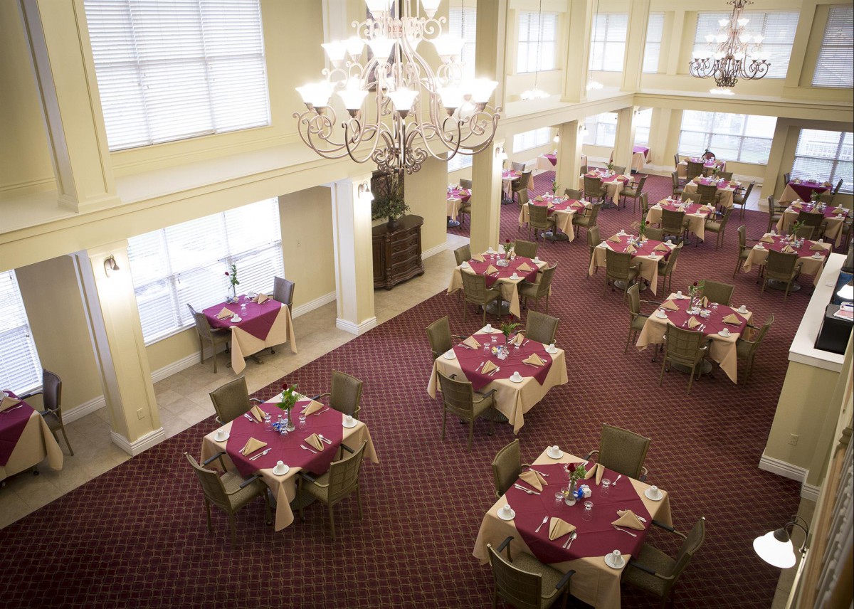 An elegant dining area featuring multiple tables with red tablecloths, surrounded by chairs, under a large chandelier.