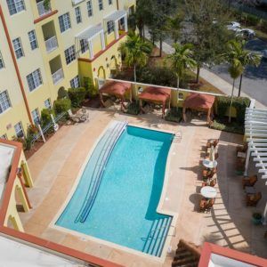 Aerial view of a hotel swimming pool surrounded by palm trees and outdoor seating on a sunny day.