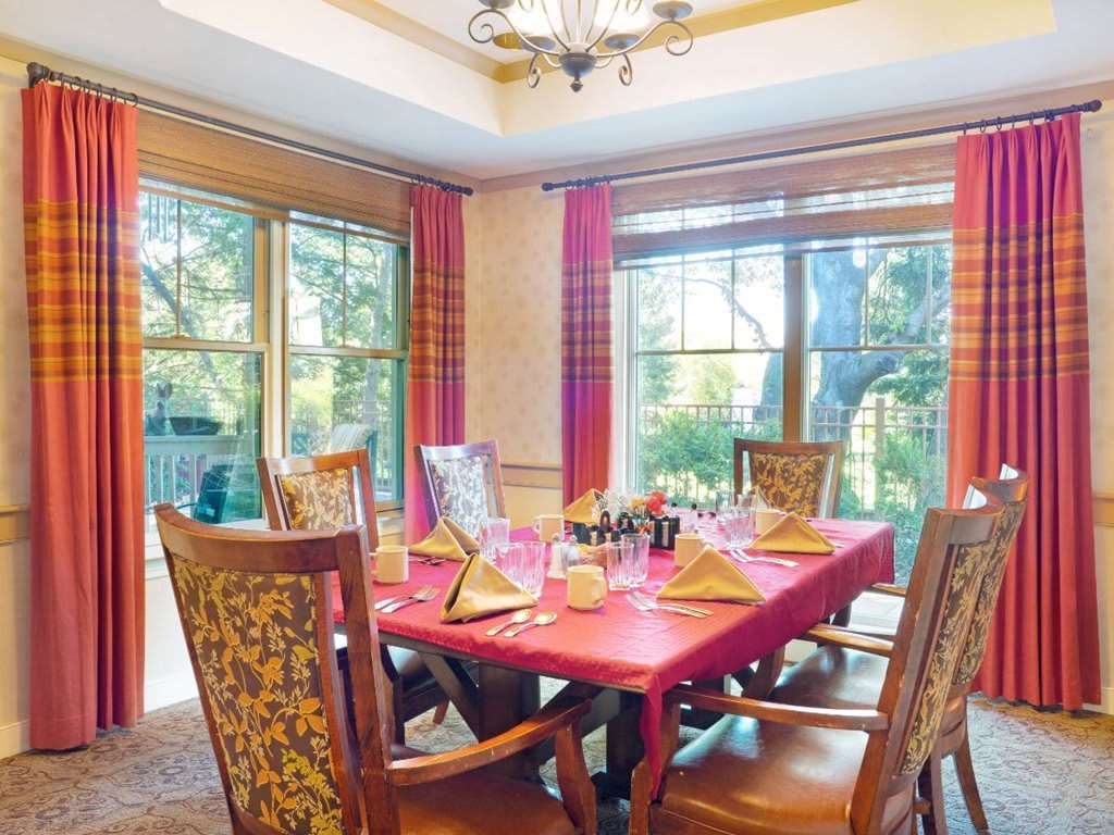 A warmly lit dining room with a red tablecloth and ornate chairs, surrounded by large windows that let in natural light.