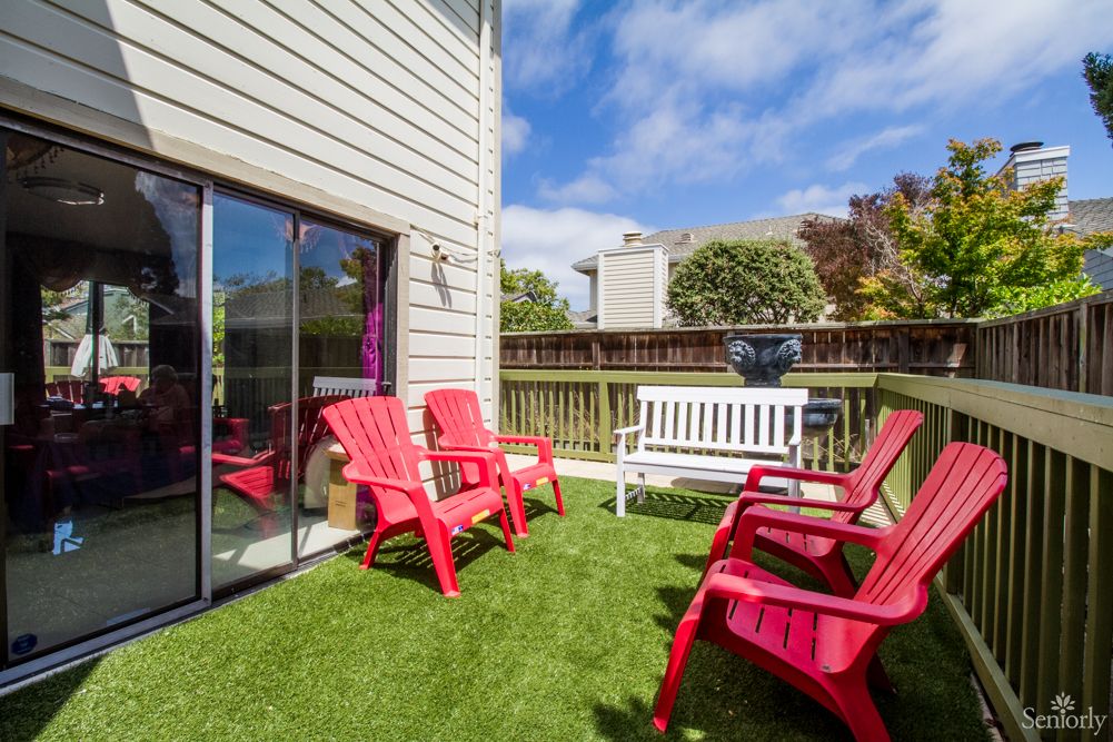 A sunny backyard patio with red chairs and a white bench on green artificial grass.