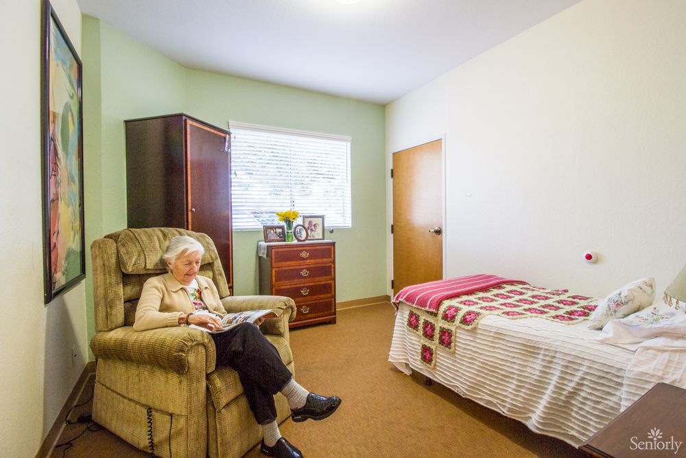 An elderly woman sits in a cozy chair reading, in a well-lit bedroom.