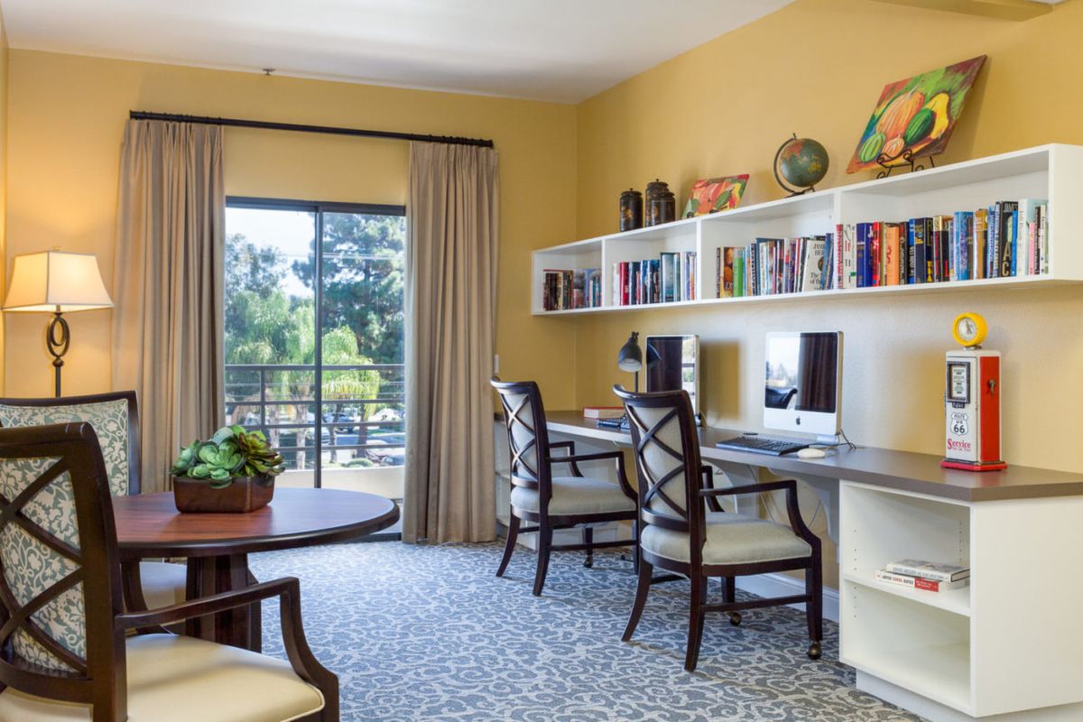 A cozy home office with dual computer workstations, bookshelves, and a view outside.