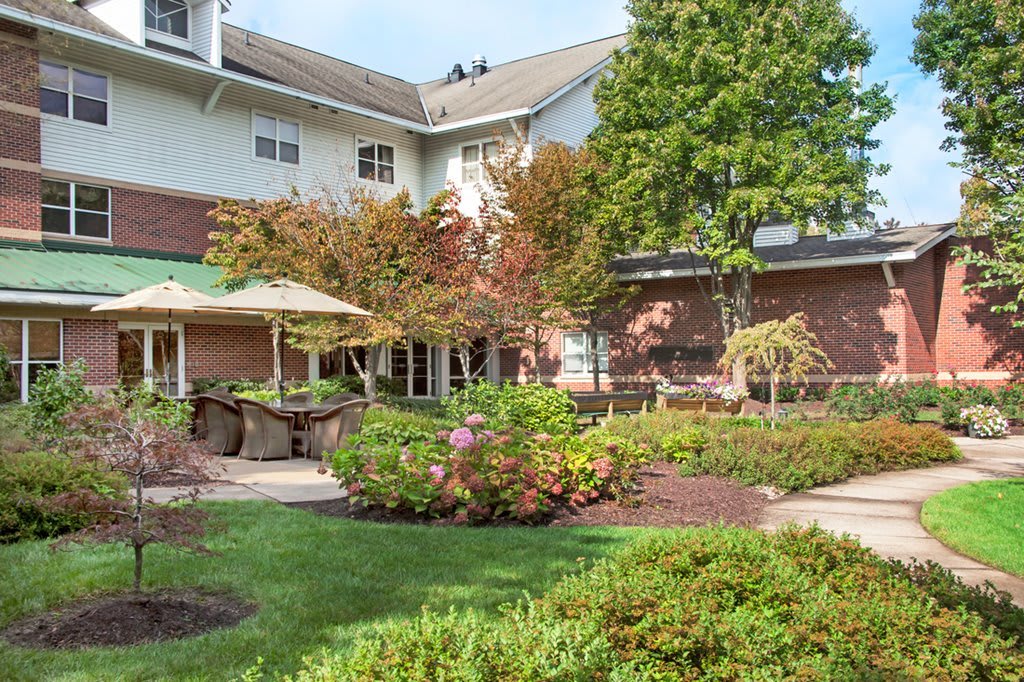 A landscaped garden area with a building in the background featuring trees, shrubs, and a patio with umbrellas.