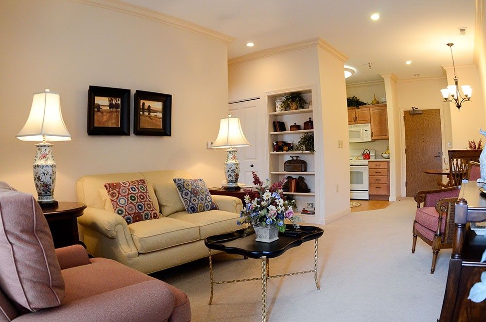 A cozy living room featuring a comfortable sofa, decorative pillows, and a coffee table with floral arrangements. The space is warmly lit by a lamp, with artwork on the walls and bookshelves leading to an adjoining kitchen.