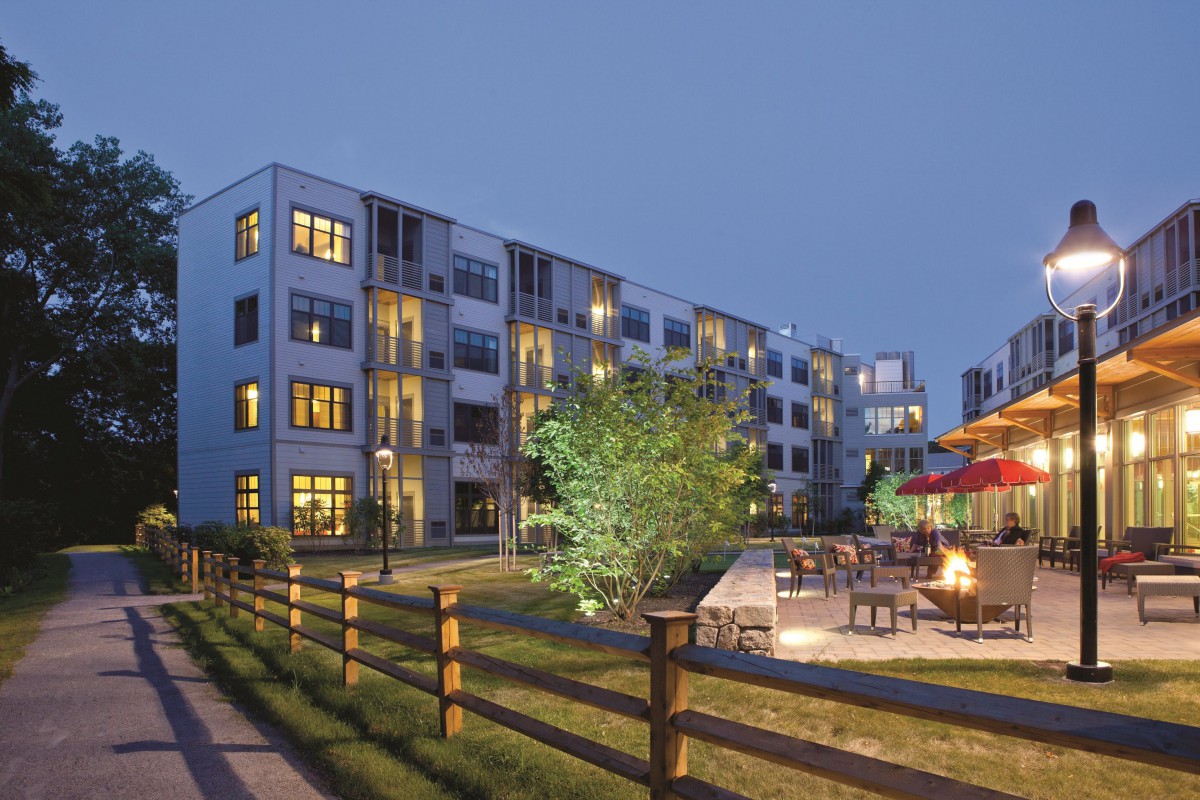 An illuminated apartment building with a fenced yard and outdoor seating area in the evening.