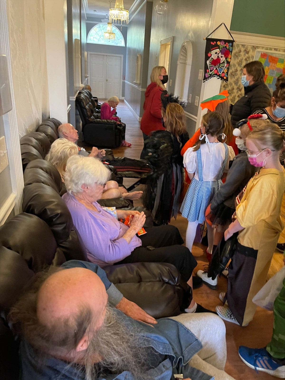 Children in costumes visit with elderly people during a social gathering indoors.