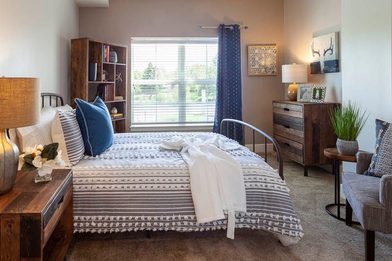 A cozy bedroom with a neatly made bed, bookshelves, and natural light coming through a large window.