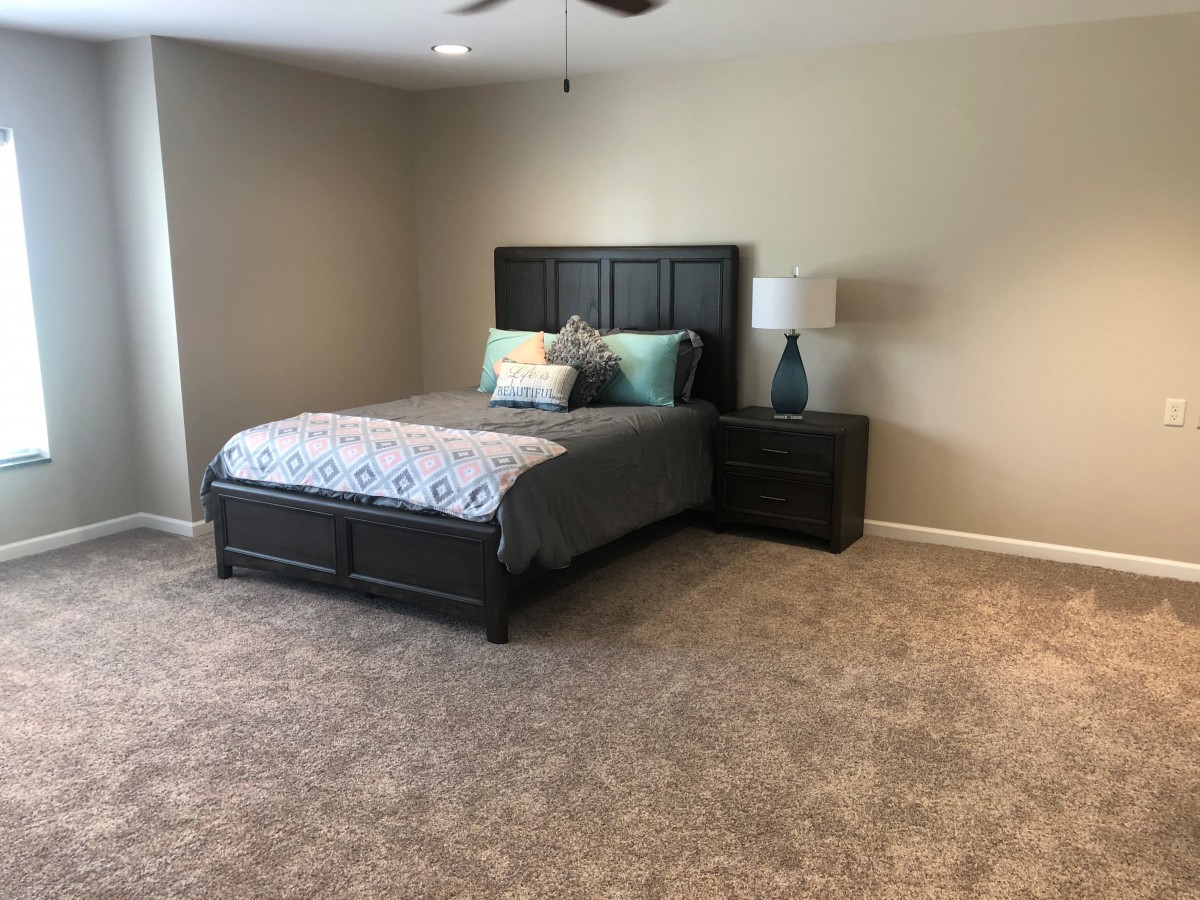 A modern bedroom with a neatly made bed, nightstand, and lamp on a carpeted floor.