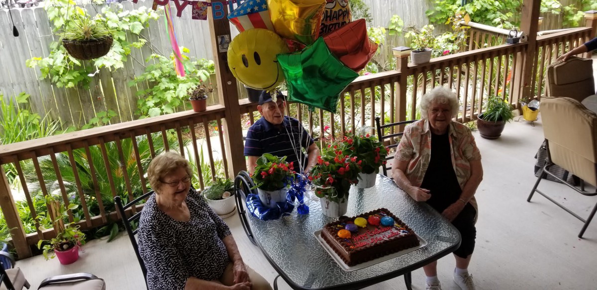 A group of people celebrating a birthday outdoors with balloons and a cake on the table.