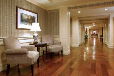 A well-lit hotel hallway with wooden flooring, featuring comfortable chairs, a side table with a lamp, and framed artwork on the wall.