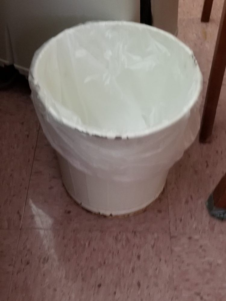 A trash can with a plastic bag liner placed on a tiled floor in an indoor setting.