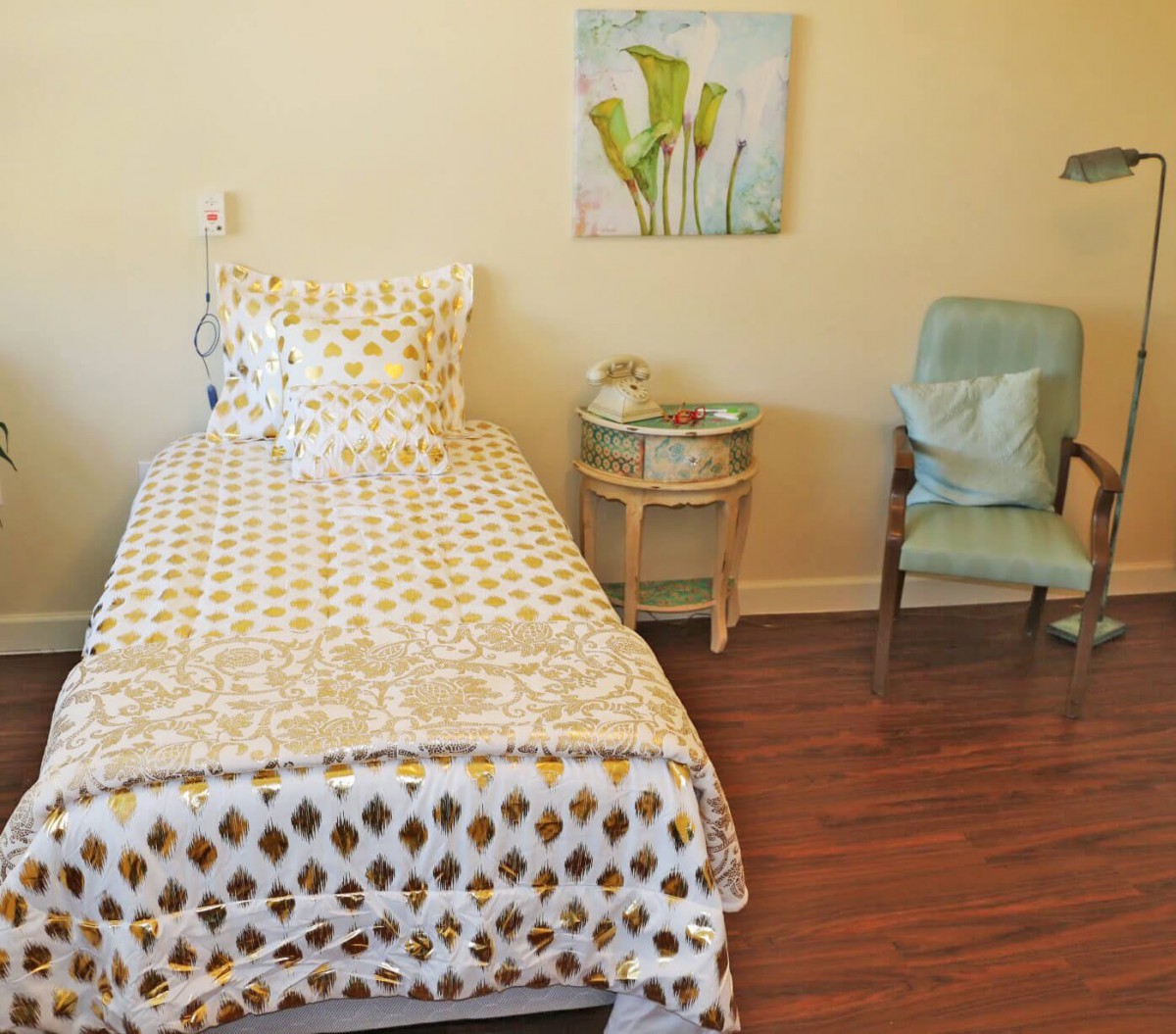 A cozy bedroom with a single bed, patterned bedding, a nightstand, and a comfortable chair on a wooden floor.