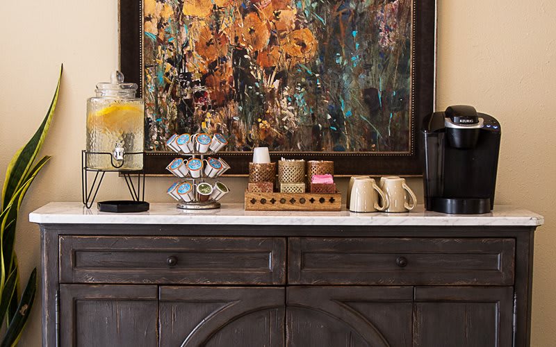 A cozy coffee station featuring a coffee maker, assorted cups, and a lemon water dispenser, all set against a colorful abstract painting.