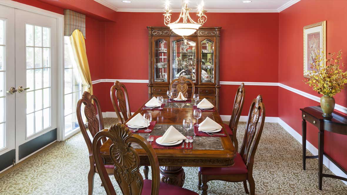A formal dining room with a wooden table and elegant decor, featuring red walls and a chandelier.