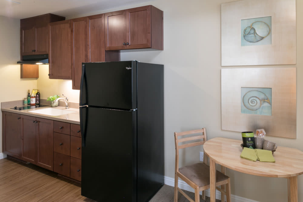A kitchen area featuring a black refrigerator, wooden cabinets, a small dining table with chairs, and framed artwork on the wall.