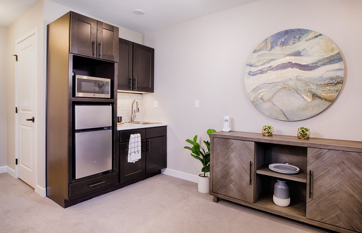 A modern kitchenette with dark cabinets, a microwave, small fridge, and minimalist decor.