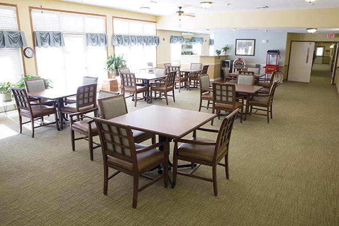 A spacious dining area with multiple tables and chairs arranged on a carpeted floor, well-lit by natural light.