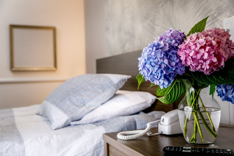 A cozy bedroom scene with a well-made bed, featuring fluffy pillows and a stylish arrangement of purple and pink hydrangeas in a vase on the nightstand, accompanied by a classic telephone.
