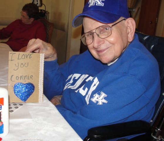 An elderly man wearing a blue hat and sweater is smiling while holding a card.