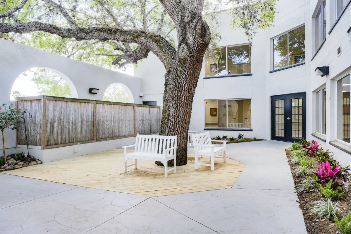 A tranquil courtyard featuring a large tree surrounded by white benches, adjacent to a building with windows and a wooden fence.