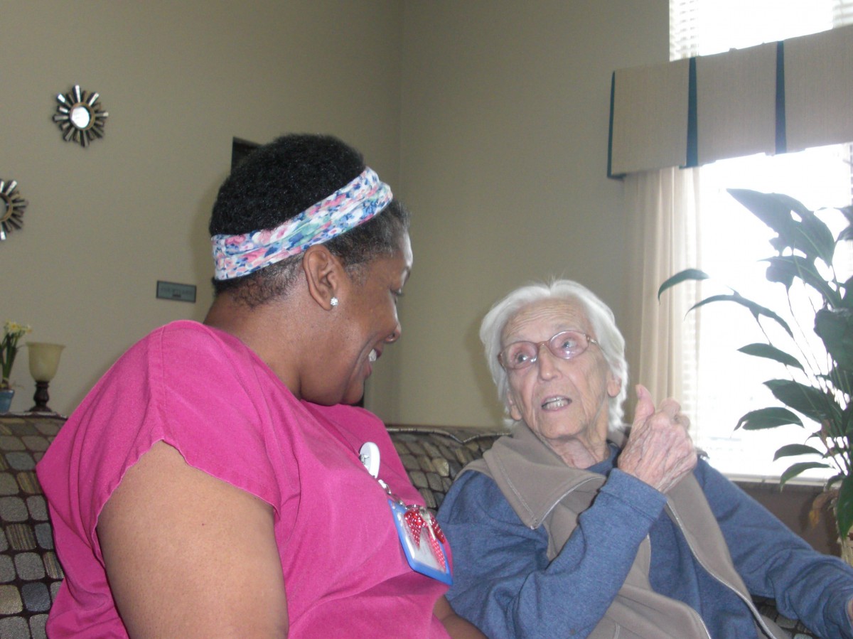 A caregiver in a pink uniform is having a conversation with an elderly woman in a cozy setting.