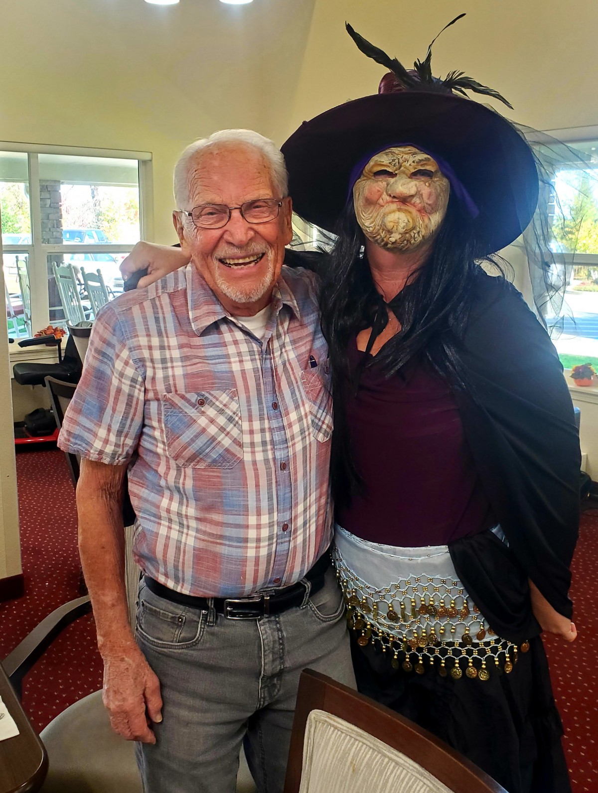 An elderly man smiles while standing next to a woman dressed in a witch costume indoors.