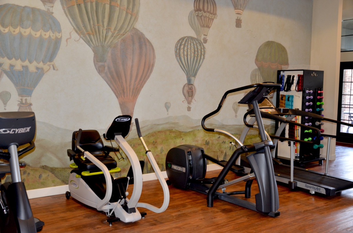 A fitness room features a stationary bike and an elliptical trainer set against a hot air balloon mural on the wall.