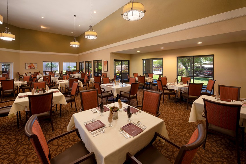 A spacious and elegantly decorated dining room with multiple tables set for guests, featuring warm lighting and large windows overlooking the garden.