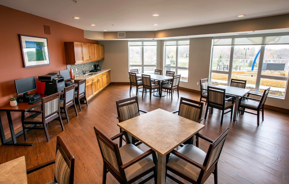 A modern dining room with tables and chairs, featuring large windows and a kitchen area.