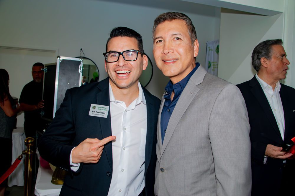 Two men in suits smiling, one wearing glasses and pointing at the other.