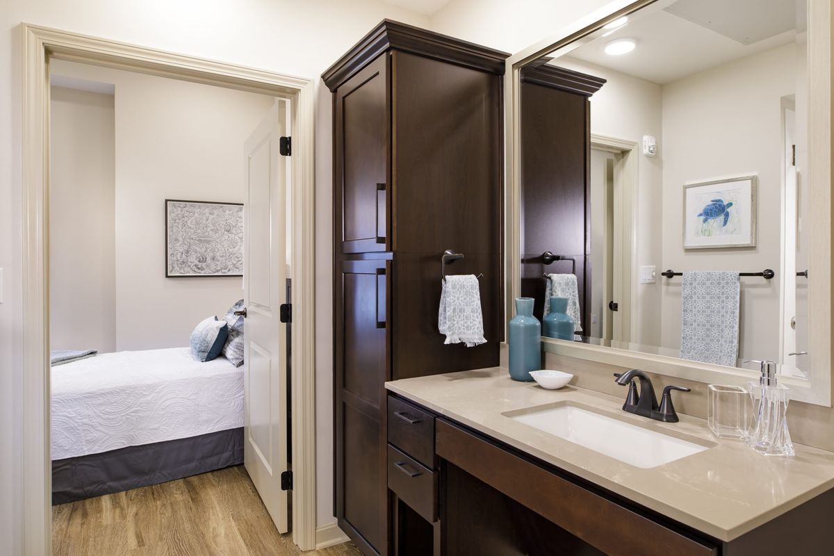 A modern bathroom with a large mirror, sink, and dark wooden cabinetry adjacent to a bedroom.