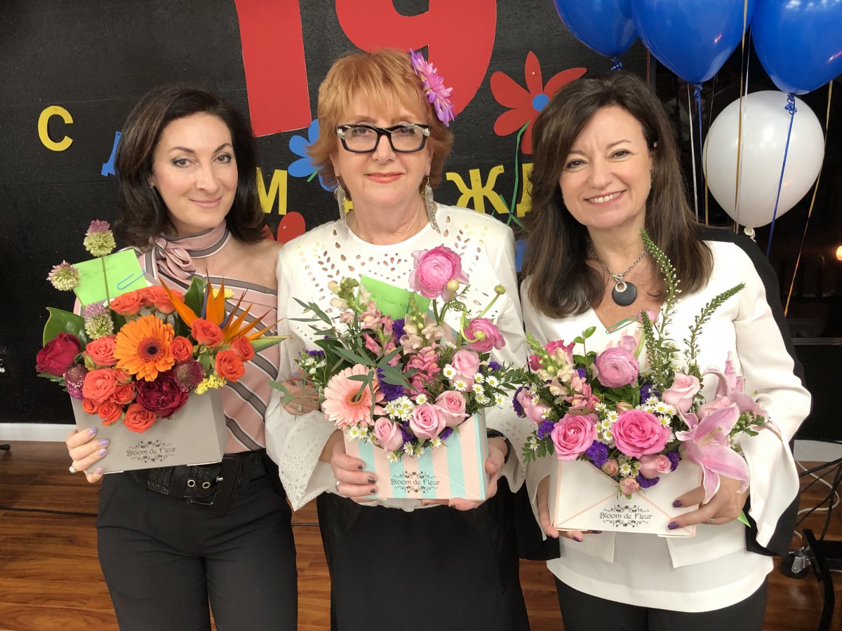Three women smiling and holding floral arrangements at a celebratory event.