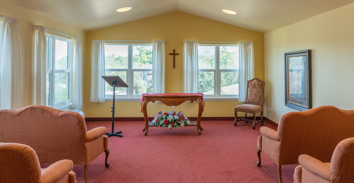 A small chapel interior with an altar and cross, featuring elegant furniture and bright windows.