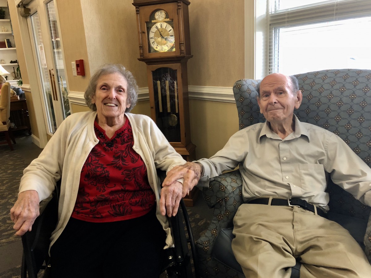 An elderly couple sitting together in a cozy living room, holding hands and smiling.